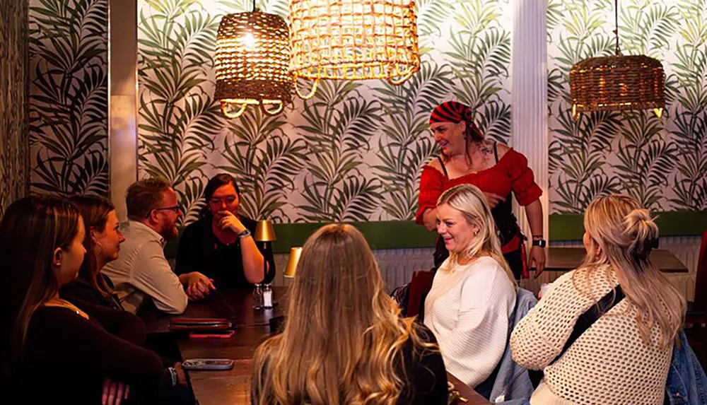 A group of people are gathered in a warmly-lit restaurant with tropical wallpaper engaging in conversation while a person in a red outfit appears to be entertaining them