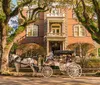 A horse-drawn carriage with a driver and passenger is parked in front of a large traditional brick house with a woman standing on the porch