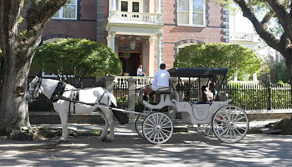 A horse-drawn carriage with a driver and passenger is parked in front of a large traditional brick house with a woman standing on the porch