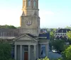 The image shows a classic church with a tall steeple and clock surrounded by lush trees with a street view and buildings in the background under a clear blue sky