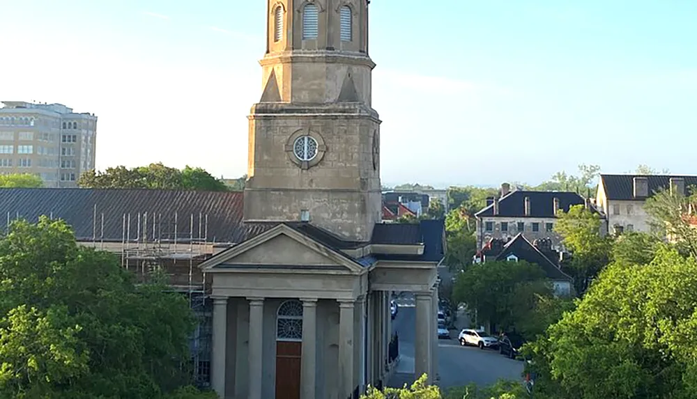 The image shows a classic church with a tall steeple and clock surrounded by lush trees with a street view and buildings in the background under a clear blue sky