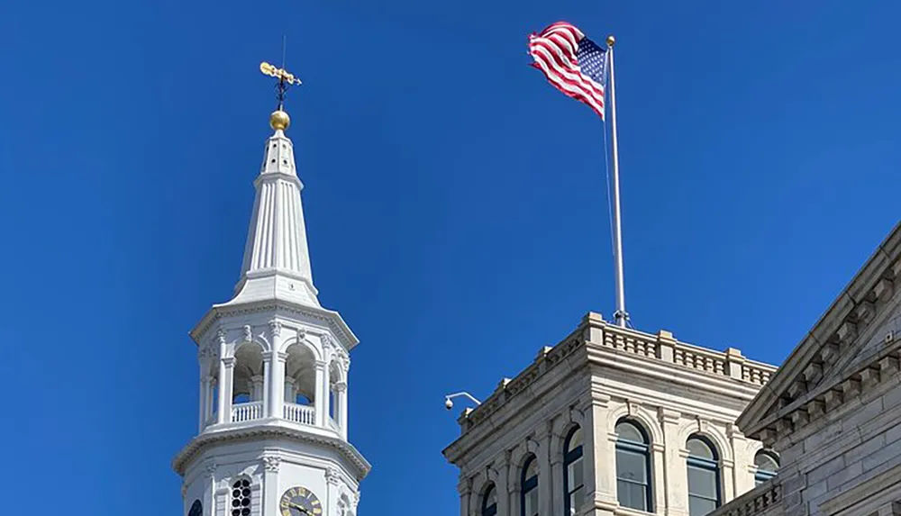 The image shows a blue sky backdrop featuring an American flag waving in the wind next to the ornate white clock tower of a historic building