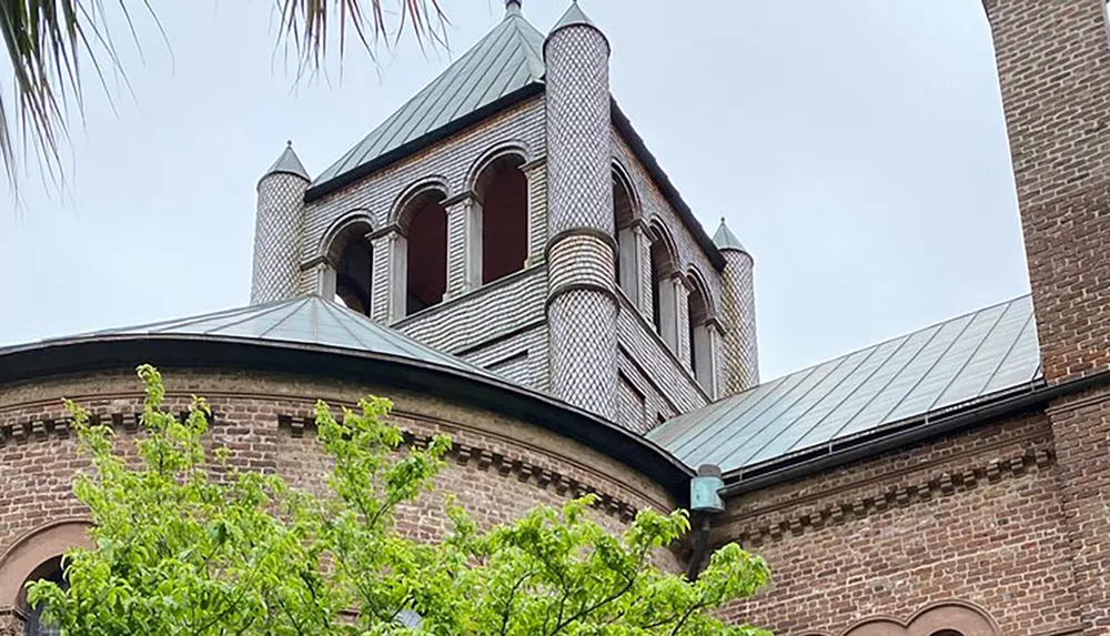 The image shows a section of a brick building with distinctive architectural elements including patterned cylindrical towers and arched windows set against a cloudy sky with foliage in the foreground