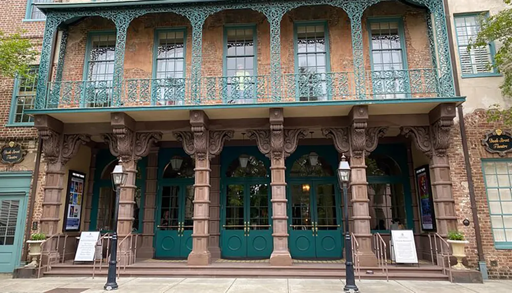 The image displays a two-story building with a unique teal wrought iron balcony and large supporting columns reminiscent of historic architectural styles