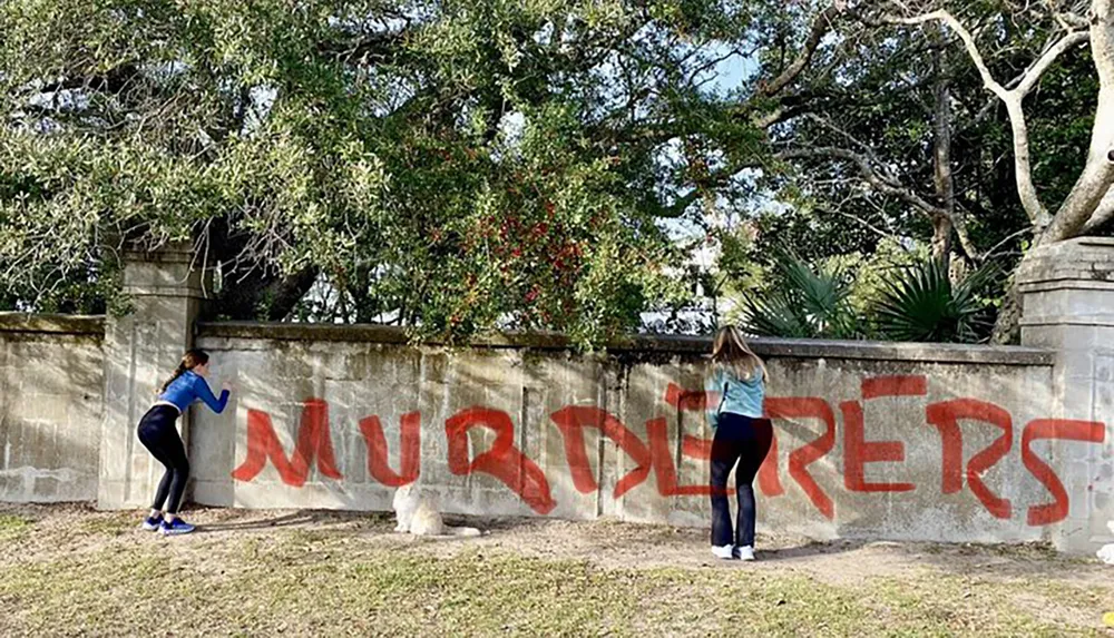 Two individuals are pictured painting over the word MURDERERS that has been graffitied on a wall