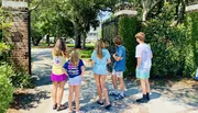 A group of children is standing at a gate entrance, looking towards a house in the distance.