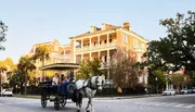 A horse-drawn carriage with passengers is moving along a street in front of a large, traditional brick building with balconies, possibly in a historic district.