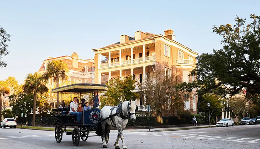 A horse-drawn carriage with passengers is moving along a street in front of a large traditional brick building with balconies possibly in a historic district