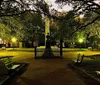 The image shows a serene night-time scene in a park with benches glowing street lamps and a statue in the center framed by the dark silhouettes of surrounding trees