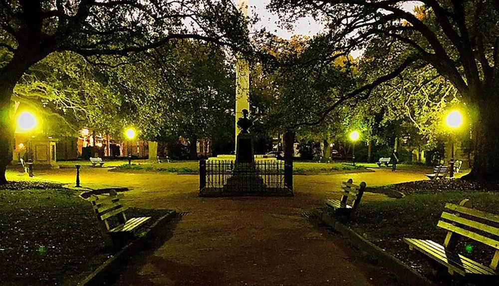 The image shows a serene night-time scene in a park with benches glowing street lamps and a statue in the center framed by the dark silhouettes of surrounding trees