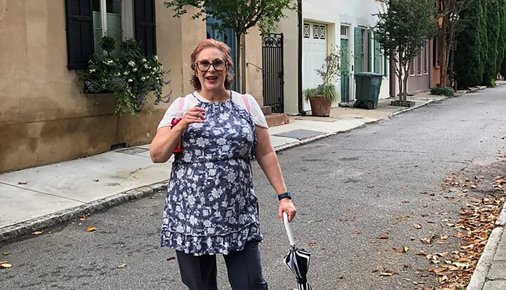 A woman in a floral dress is standing on a quiet street with a folding chair in her hand smiling at the camera