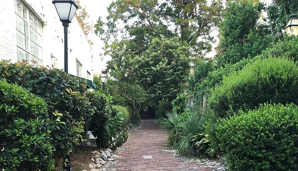 A serene brick pathway flanked by lush greenery and street lamps leads through a quiet garden