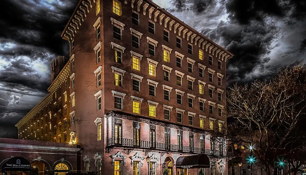 The image presents an atmospheric night view of a historic multi-story building with warm lights shining through the windows against a dramatic cloudy sky