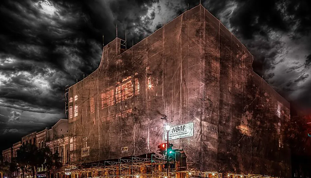 The image depicts a large ominous-looking building shrouded in a protective mesh under a brooding cloud-filled sky at dusk or nighttime with some illuminated windows and urban infrastructure visible