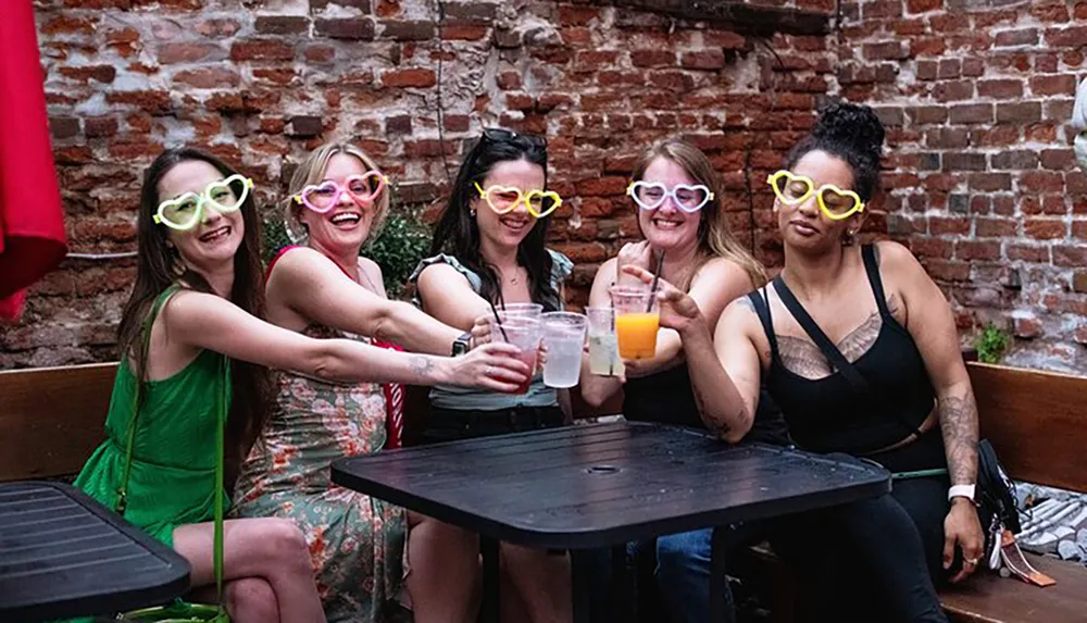 A group of five women wearing oversized heart-shaped sunglasses are enjoying drinks and smiling together at an outdoor table