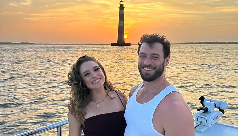 A smiling couple pose for a photo on a boat with a sunset behind a lighthouse in the background