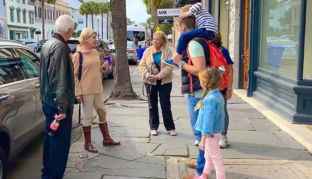 A group of people including a family with children and a man carrying a young child on his shoulders appears to be engaged in conversation on a city sidewalk lined with parked cars and palm trees