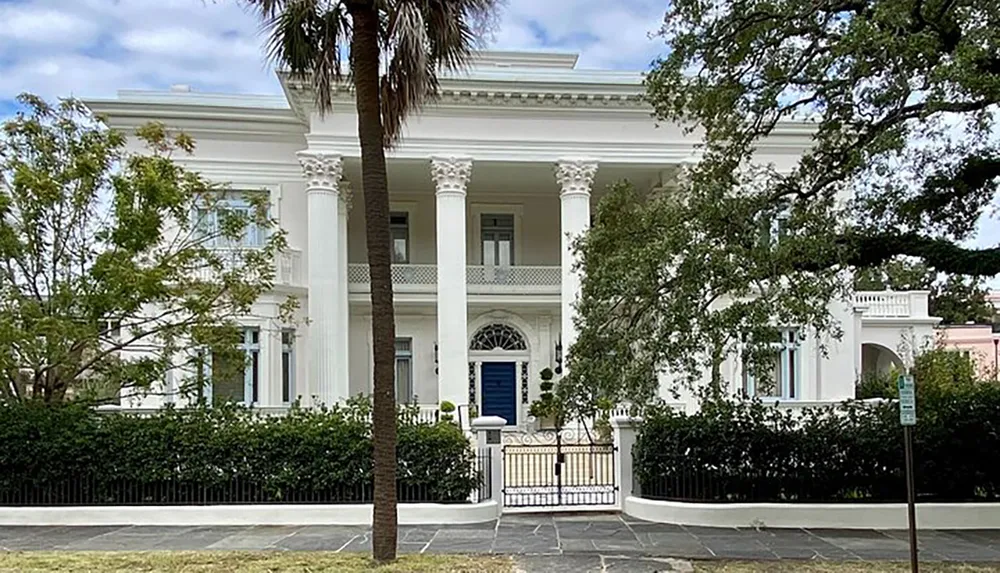 This image shows a large stately white house with tall columns a balcony and lush greenery reflecting a classic and elegant architectural style