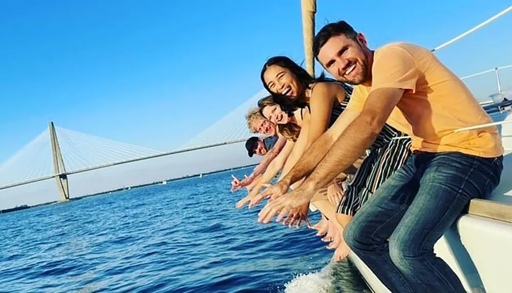 A group of smiling people are sitting on the edge of a sailboat extending their hands over the water against the backdrop of a clear sky and a suspension bridge