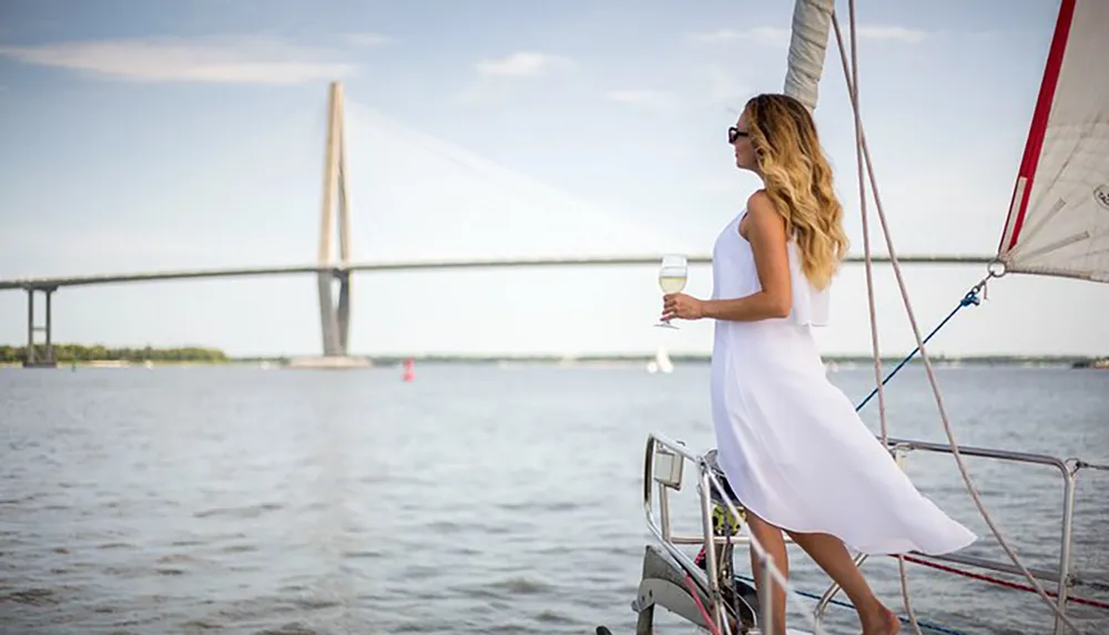 A woman in a white dress enjoys a glass of wine on a sailboat with a large bridge in the background