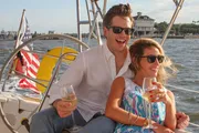 A happy couple is enjoying a sunny day on a sailboat, each holding a glass of wine, with the American flag in the background.