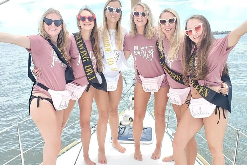 A group of women are smiling and posing together on a boat dressed in coordinating outfits with celebratory sashes that suggest they are part of a bachelorette party