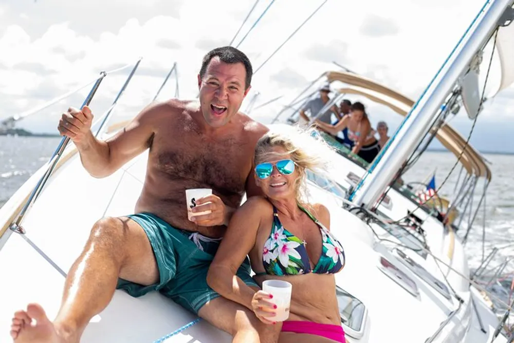 A man and a woman are enjoying themselves on a sailing boat with the man making a joyful expression while holding a drink and the woman smiling at the camera both in swimwear with other people in the background