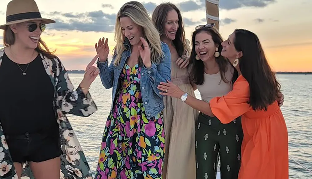 A group of five joyful women are sharing a moment of laughter and celebration on a boat with a beautiful sunset in the background