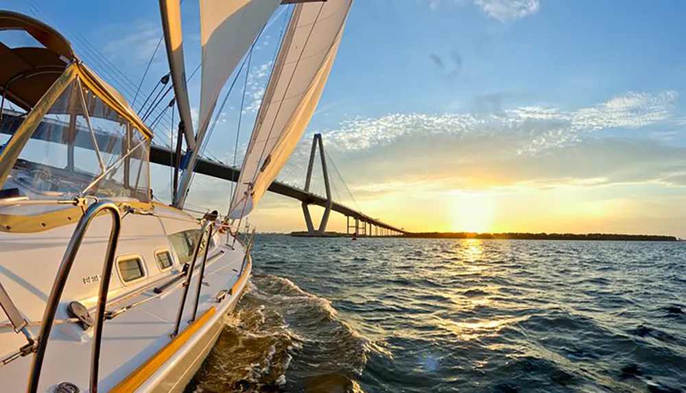 A sailboat is sailing near a large bridge at sunset with the sunlight reflecting off the waters surface