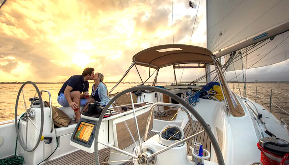 Two people are sharing a kiss on the deck of a sailboat with a beautiful sunset in the background