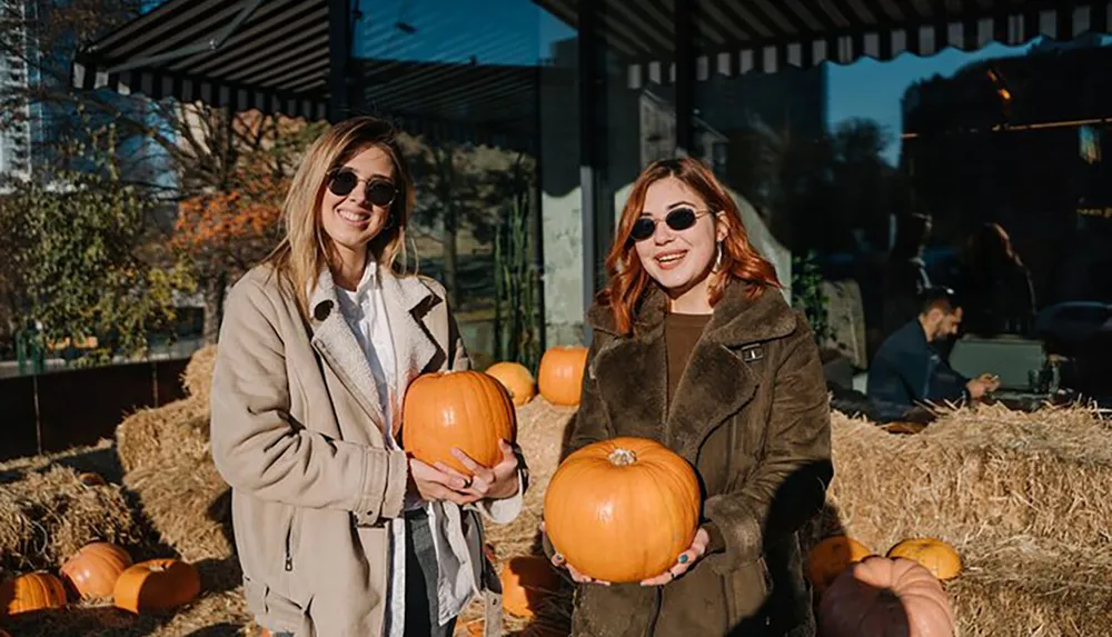 Two smiling individuals wearing sunglasses are holding pumpkins in front of a haystack possibly at a pumpkin patch or a fall festival setting