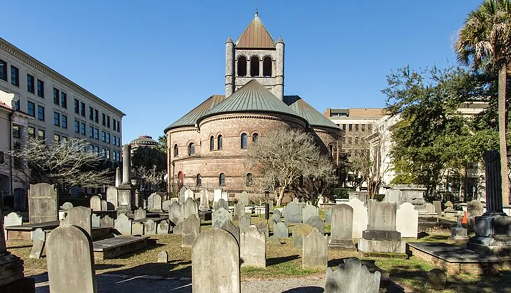 The image shows a historic church with a distinct round architecture and bell tower overlooking an old cemetery with numerous gravestones contrasting with the modern buildings in the background on a sunny day