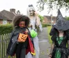 Three individuals in Halloween costumes are trick-or-treating with one dressed as a devil another as a ghost or zombie and the third as a witch walking along a residential sidewalk