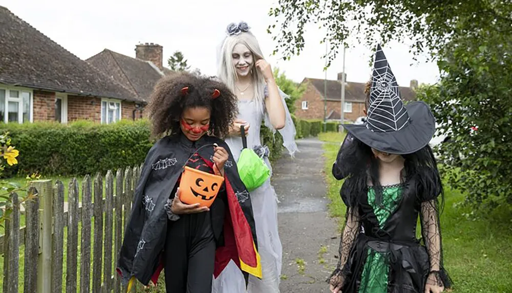 Three individuals in Halloween costumes are trick-or-treating with one dressed as a devil another as a ghost or zombie and the third as a witch walking along a residential sidewalk