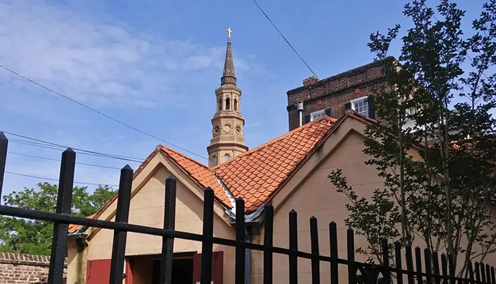 The image shows a view of the top of an old church with a tall steeple behind a smaller building with a terracotta tile roof all enclosed by a black metal fence with trees partially obscuring the view under a partly cloudy sky