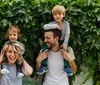 A happy family with two young children on their parents shoulders is posing in front of a leafy green backdrop