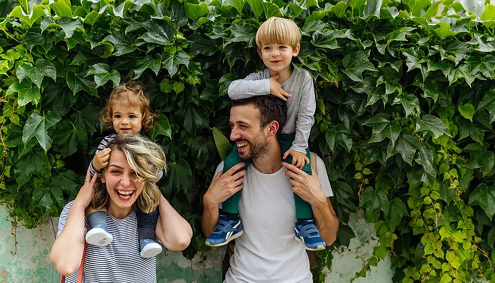 A happy family with two young children on their parents shoulders is posing in front of a leafy green backdrop