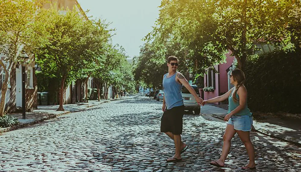 A man and a woman are holding hands while crossing a cobblestone street in a charming neighborhood with trees and colorful buildings