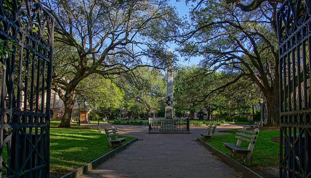 A tranquil park pathway leads to an obelisk monument framed by wrought-iron gates and shaded by mature sprawling trees