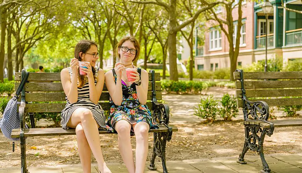 Two people are sitting on a park bench enjoying drinks on a sunny day