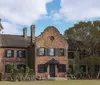 The image shows a two-story brick building with a unique architectural style featuring a distinctive central gable with ornamental detailing nestled in a landscape with mature trees and a partly cloudy sky above