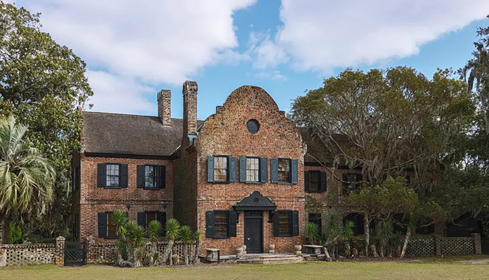 The image shows a two-story brick building with a unique architectural style featuring a distinctive central gable with ornamental detailing nestled in a landscape with mature trees and a partly cloudy sky above