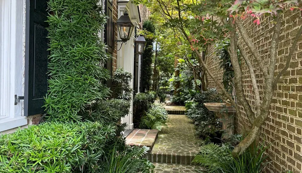 The image shows a serene and lush garden pathway flanked by brick walls and green foliage with a hint of a traditional lantern illuminating the space