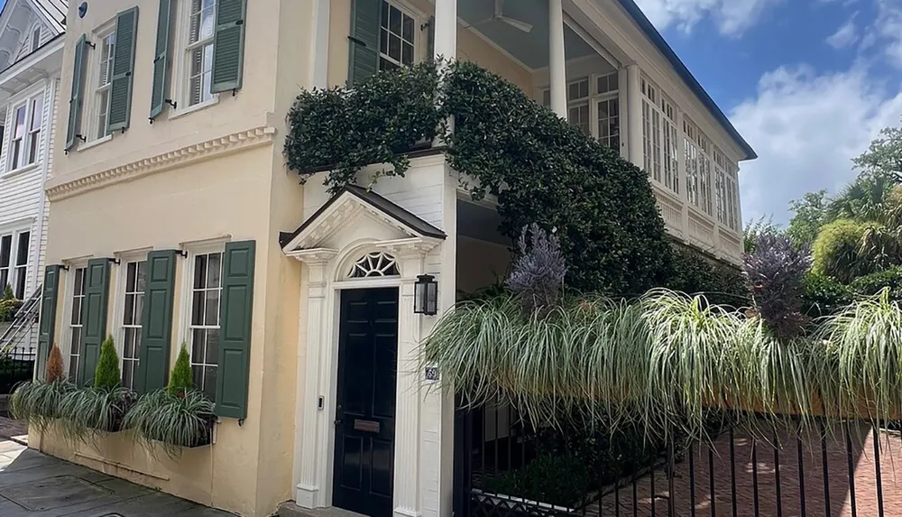 The image shows a classic two-story beige house with green shutters an elegant front door an ivy-covered wall and lush landscaping under a clear sky