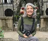 A smiling person with gray hair is sitting on a step holding a camera with greenery and an ornate metal gate in the background