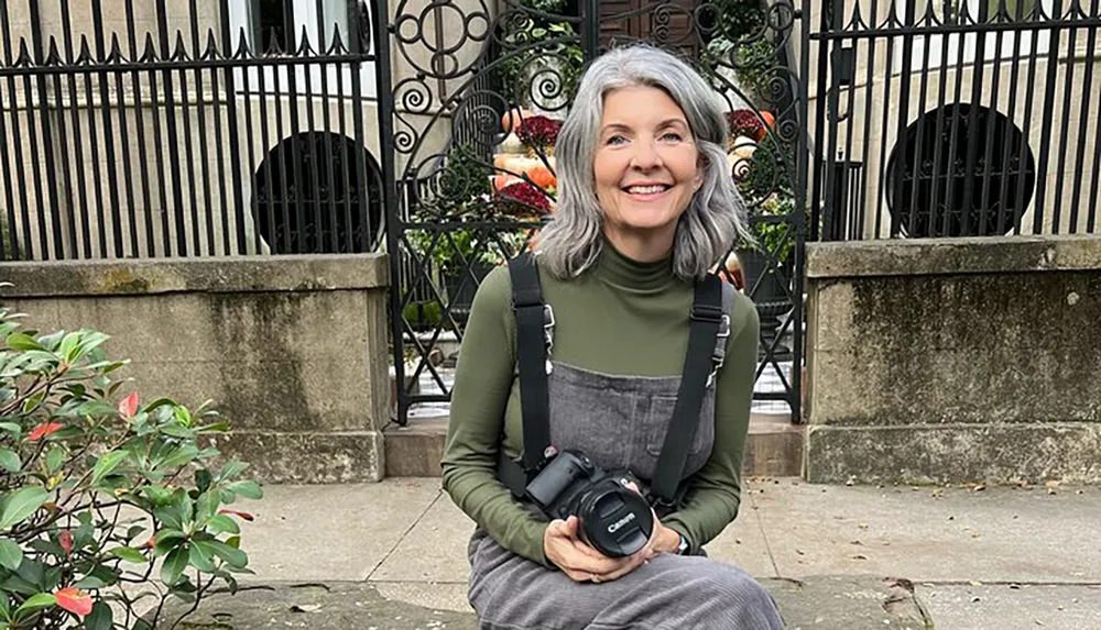 A smiling person with gray hair is sitting on a step holding a camera with greenery and an ornate metal gate in the background