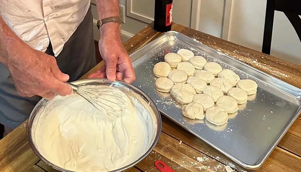 A person is preparing dough in a bowl near a baking sheet with rolled out biscuit shapes on a floured surface