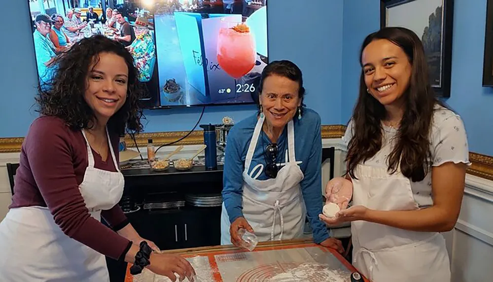 Three people wearing aprons and gloves are smiling at the camera seemingly enjoying themselves while working or participating in a cooking or confectionery activity with a TV screen and various items on the background tables