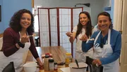 Three smiling people, likely a family, are wearing aprons and tasting food in a kitchen setting.