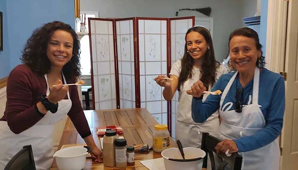 Three smiling people likely a family are wearing aprons and tasting food in a kitchen setting